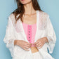 Woman wearing open front lace detail cardigan with 3/4 bell sleeves and frayed edge over pink top and white pants.