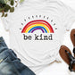 White graphic round neck short sleeve t-shirt with rainbow design and "be kind" text, styled with jeans and sandals.