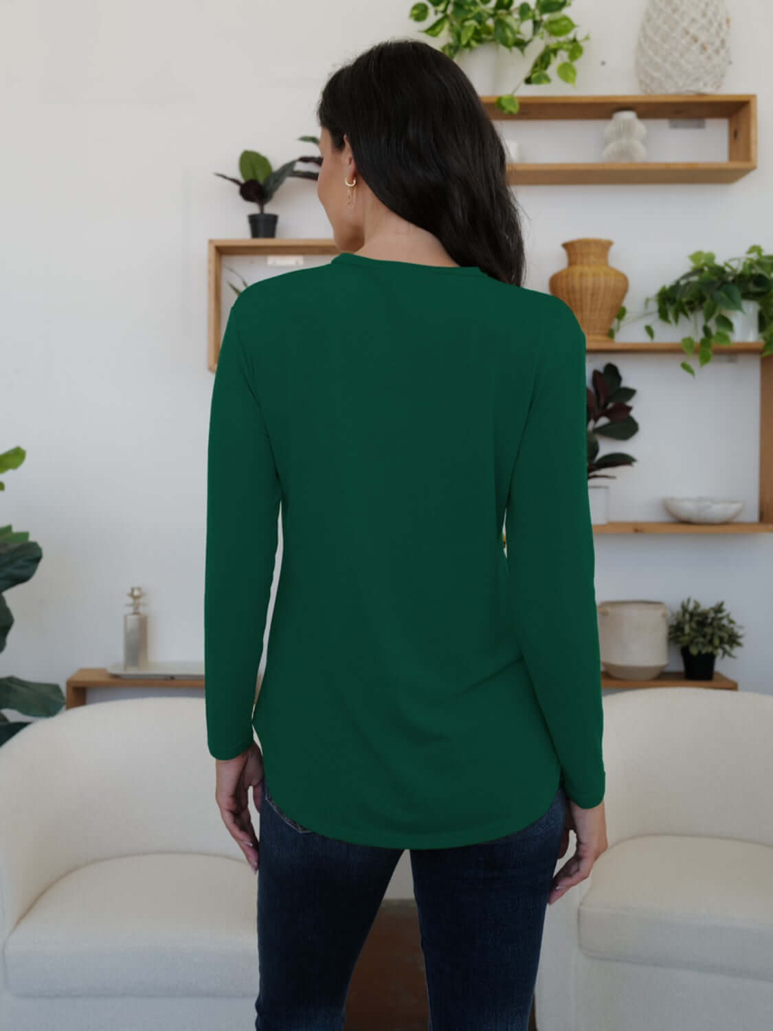 Woman wearing FAM-FAM round neck long sleeve t-shirt in green, standing with back to camera in stylish room with plants and shelves.