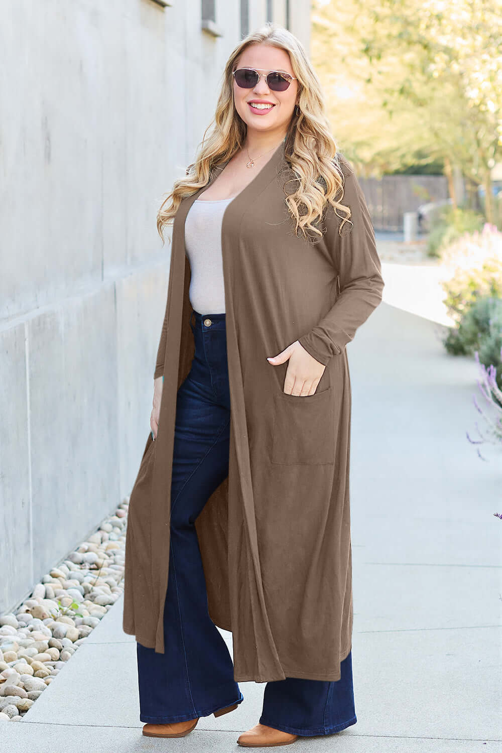 Woman wearing a brown open front long sleeve cover-up with pockets, paired with jeans and sunglasses, standing outdoors