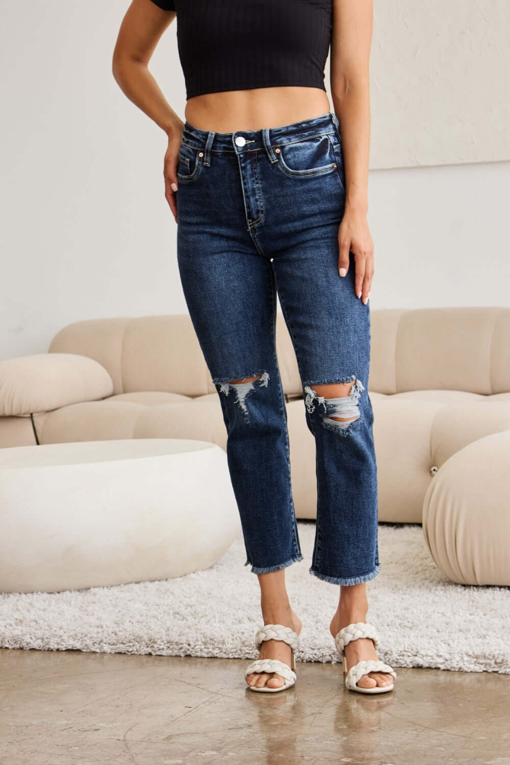 Woman wearing RFM Jeans Crop Dylan Full Size Tummy Control Distressed High Waist Raw Hem Jeans in living room.