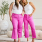 Two women wearing matching pink jeans and white tops, smiling and posing stylishly in a minimalist room setting.