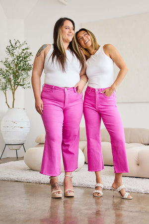 Two women wearing vibrant pink high-waisted jeans and white tank tops, standing confidently in a stylish modern living room.