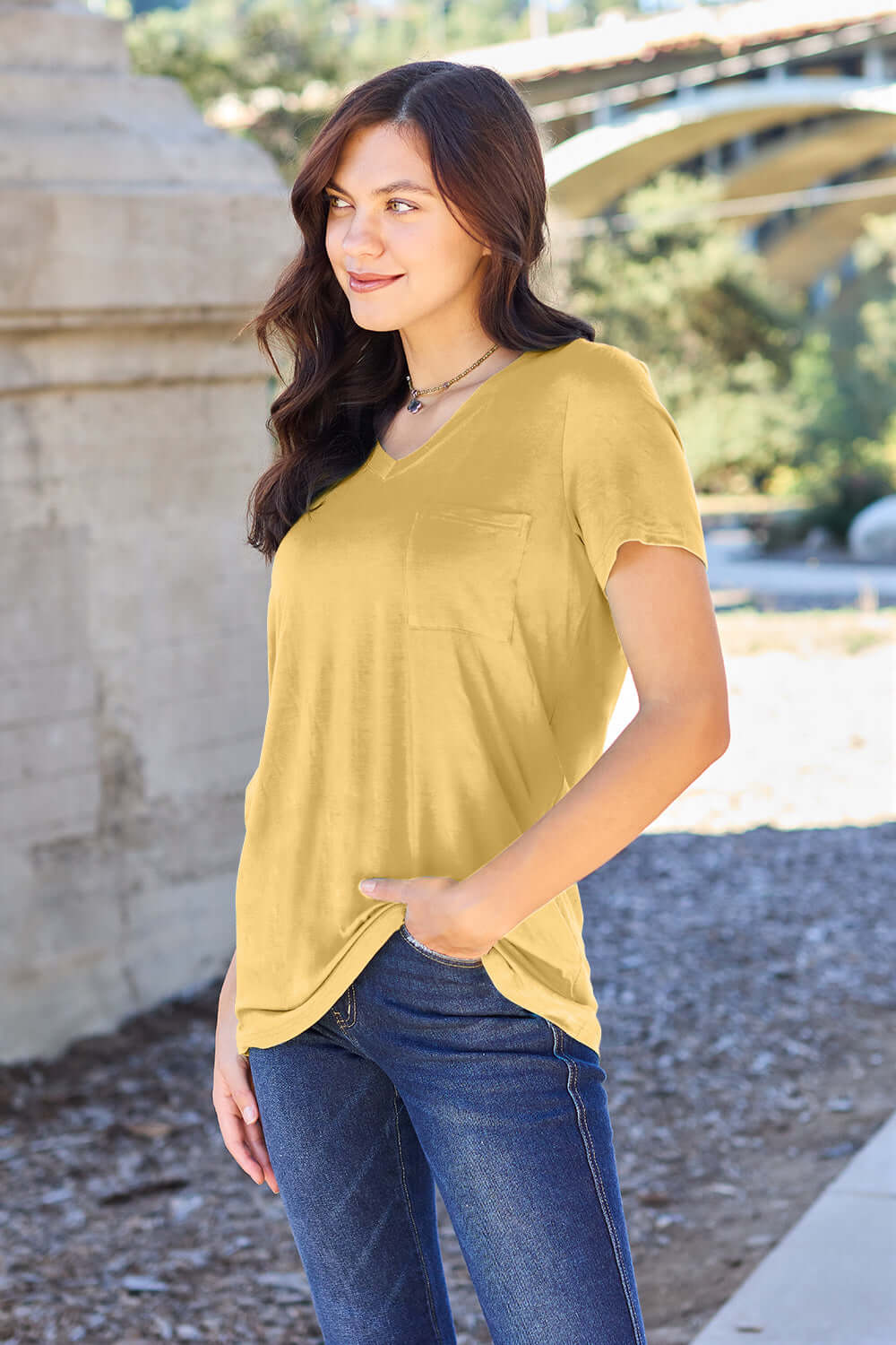 Woman wearing Basic Bae Bamboo Full Size V-Neck Short Sleeve T-Shirt in yellow, standing outdoors near a stone wall.