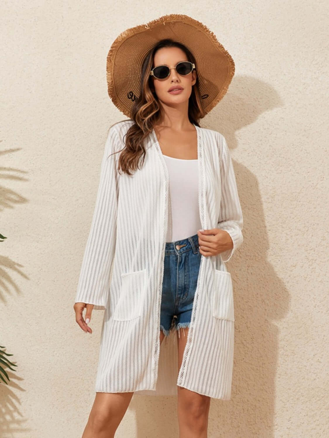 Woman wearing a lace detail open front long sleeve cover-up, white tank top, denim shorts, sunhat, and sunglasses