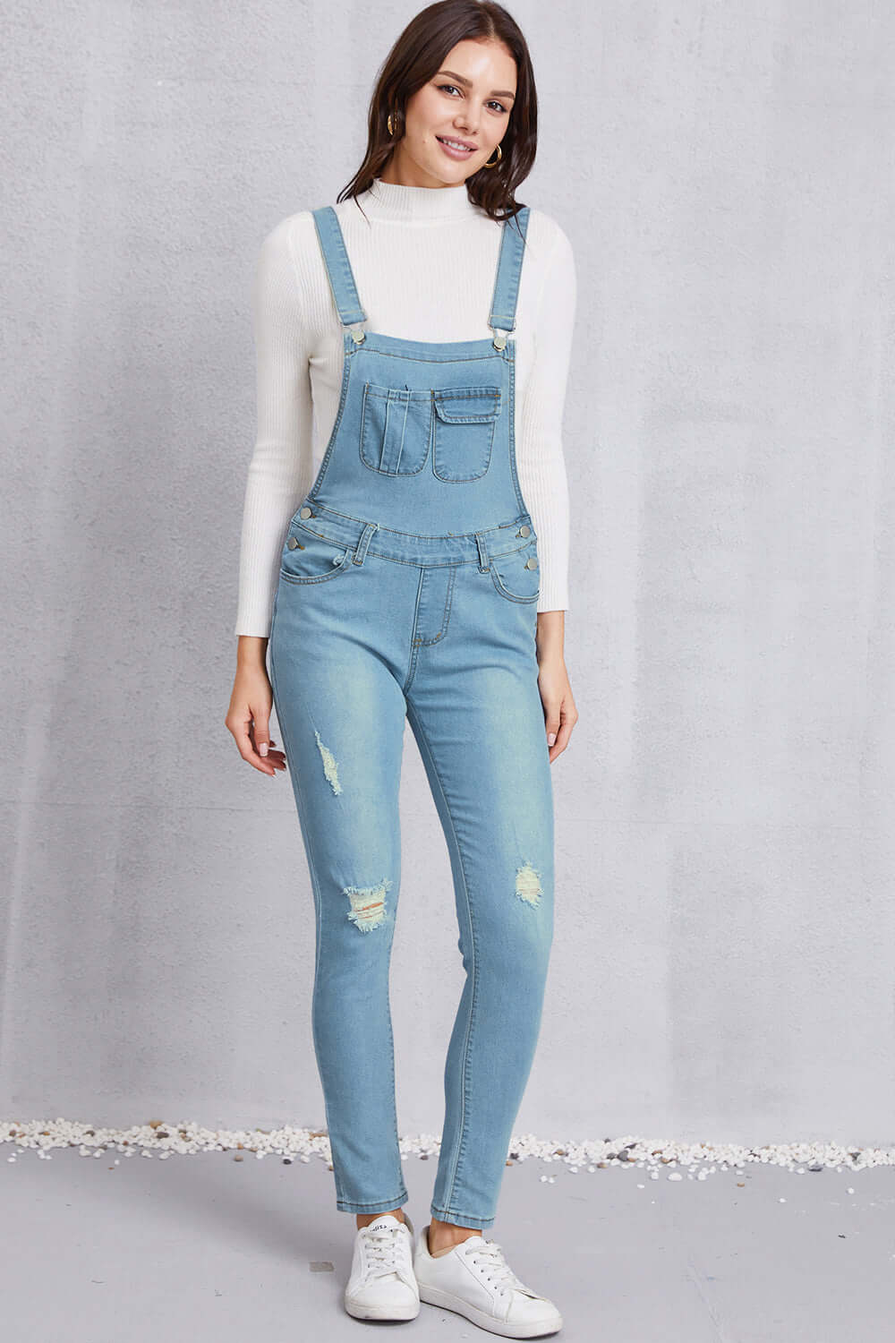 Woman wearing distressed washed denim overalls with pockets, slightly stretchy mid waist fit, made of 65% cotton and 35% polyester