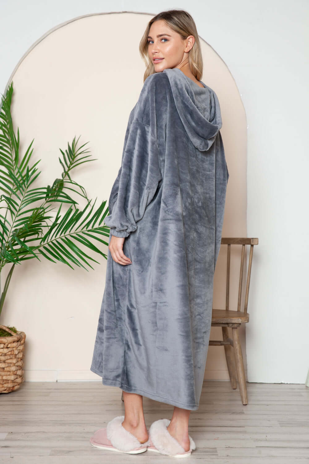 Cozy woman wearing a gray hooded midi lounge dress with pockets, standing in a stylish room with plants.