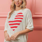 Woman wearing striped heart contrast knit top with red and white heart design on a striped background, standing against a pink wall.