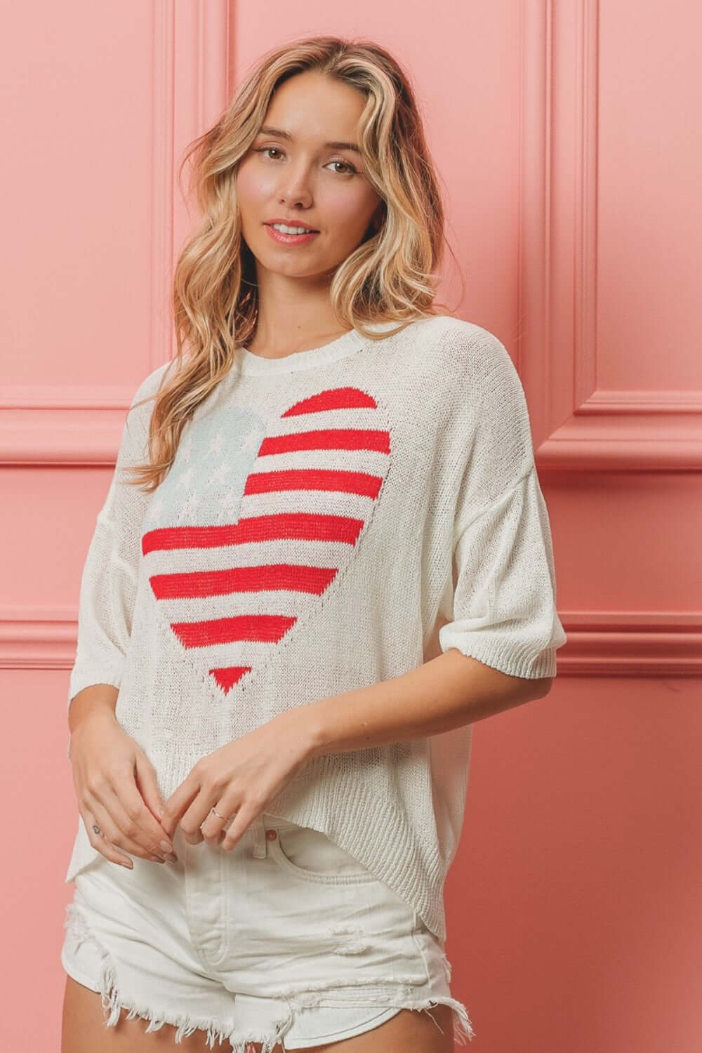 Woman wearing striped heart contrast knit top with red and white heart design on a striped background, standing against a pink wall.