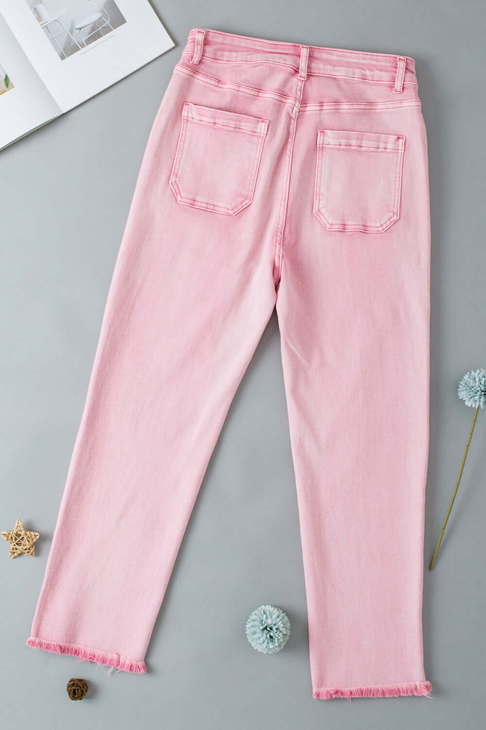 Pink raw hem button-fly jeans with back pockets for petites laid flat on a grey background.