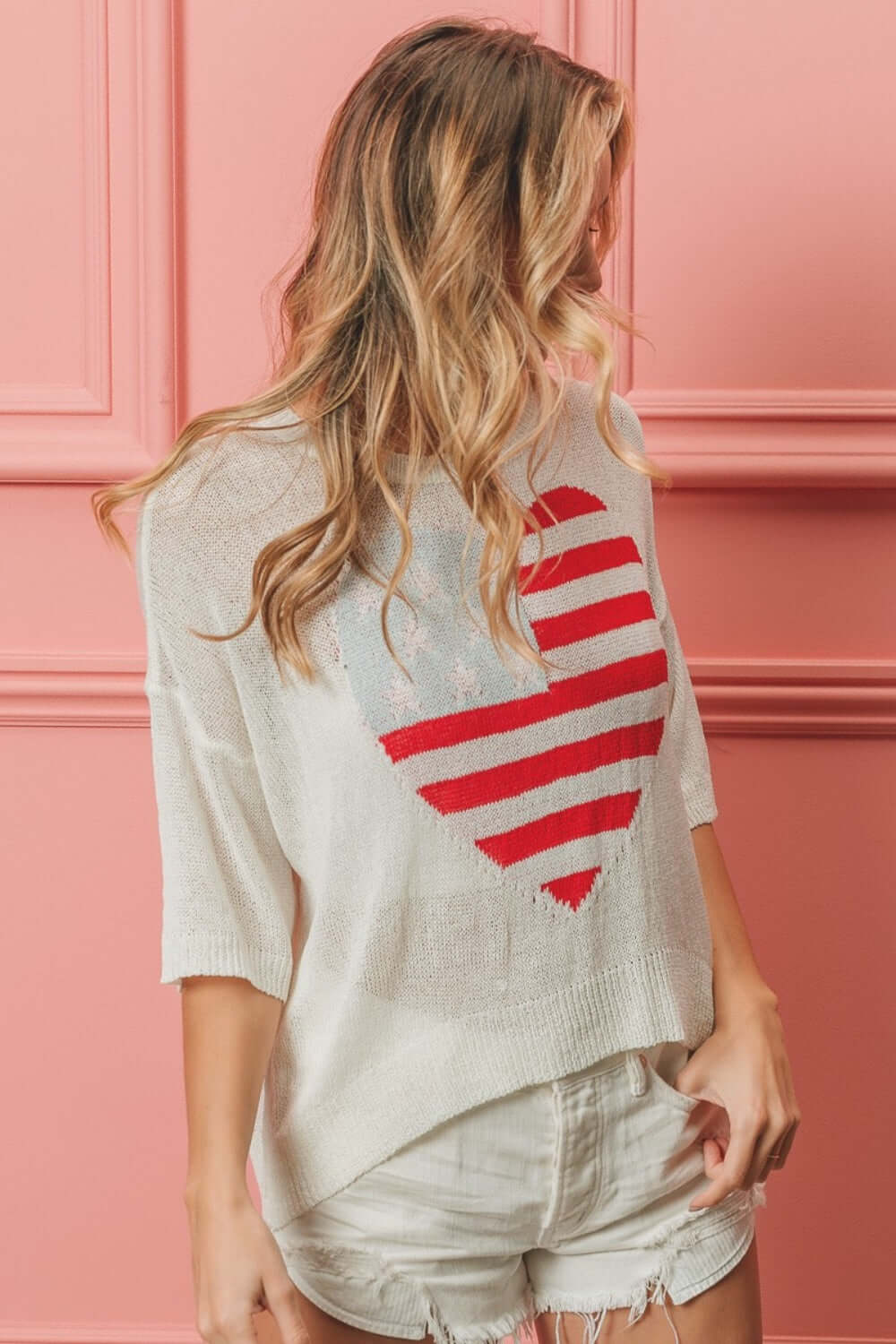 Woman wearing a white striped heart contrast knit top with red stripes on a heart design and white shorts against a pink background