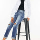 Woman wearing high waist distressed hem detail cropped straight jeans with rolled cuffs and white jacket, seated on a stool, showcasing trendy edgy style.