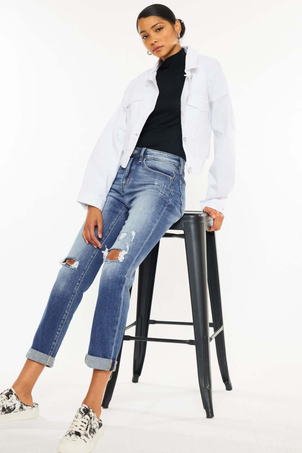 Woman wearing high waist distressed hem detail cropped straight jeans with rolled cuffs and white jacket, seated on a stool, showcasing trendy edgy style.