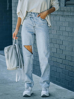 Woman wearing Distressed Asymmetric Waist Jeans with ripped details, holding a gray backpack, standing against a brick wall.