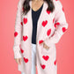 Woman wearing Bella Road Heart Open Front Long Sleeve Cardigan in pink with red hearts, standing against a coral background.