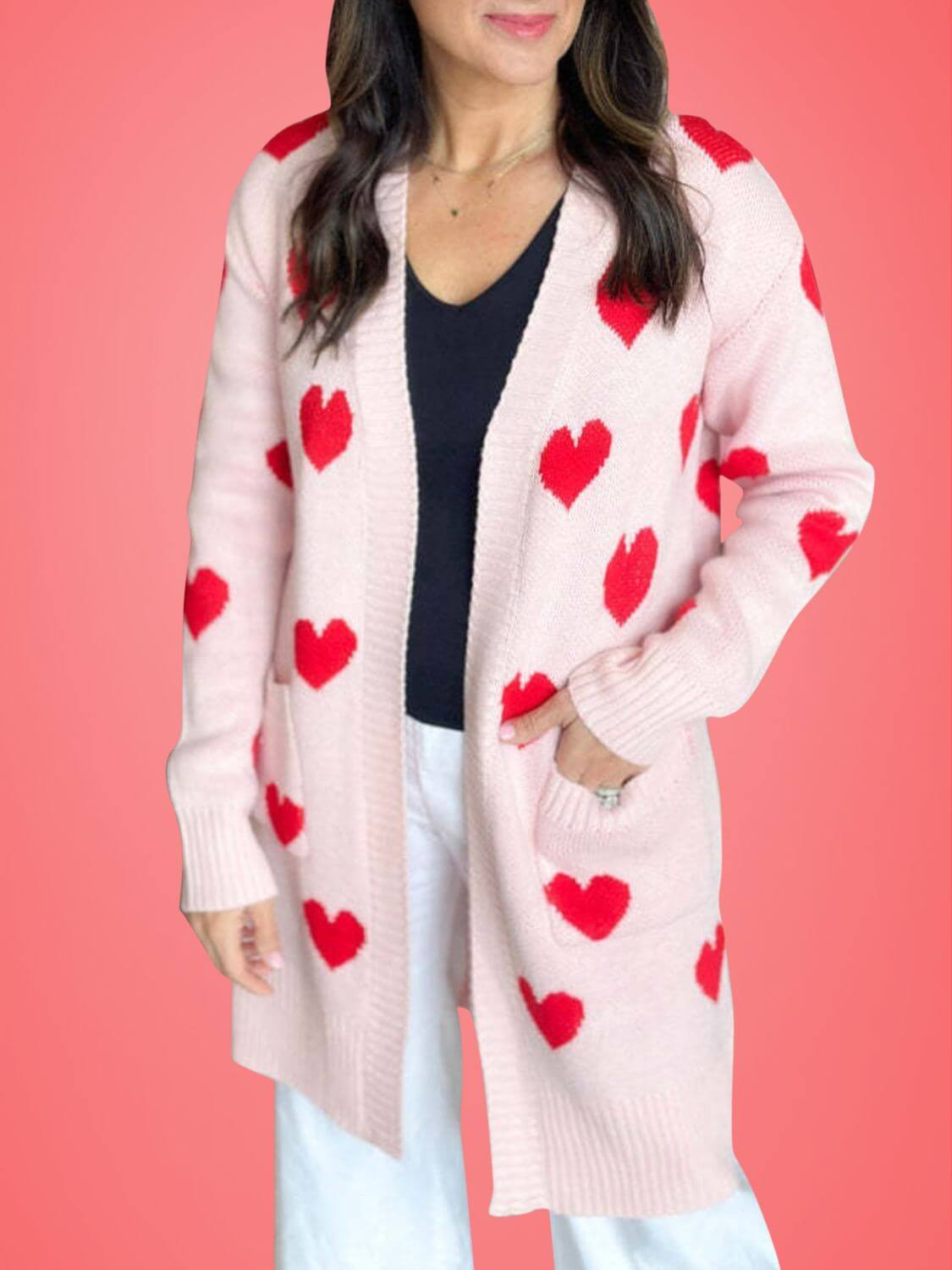 Woman wearing Bella Road Heart Open Front Long Sleeve Cardigan in pink with red hearts, standing against a coral background.