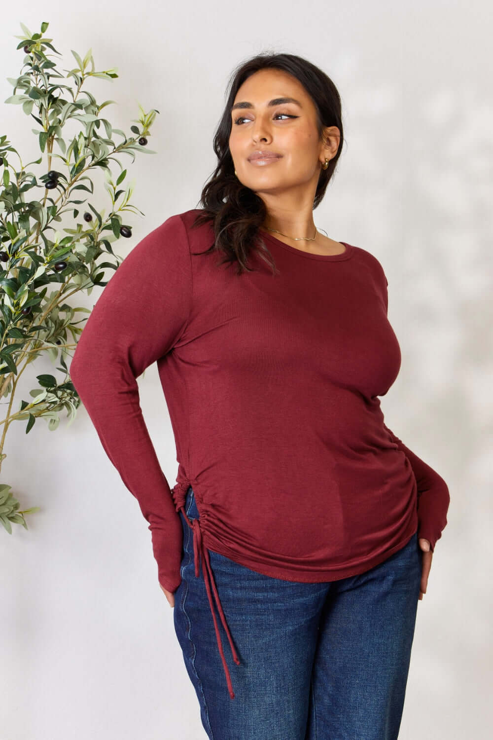 Curve model wearing drawstring round neck long sleeve top in burgundy, paired with blue jeans, standing near a plant.