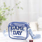 Transparent crossbody bag with "GAME DAY" letters, blue glitter trim, and zigzag strap on a white chair. Perfect for stadium events.