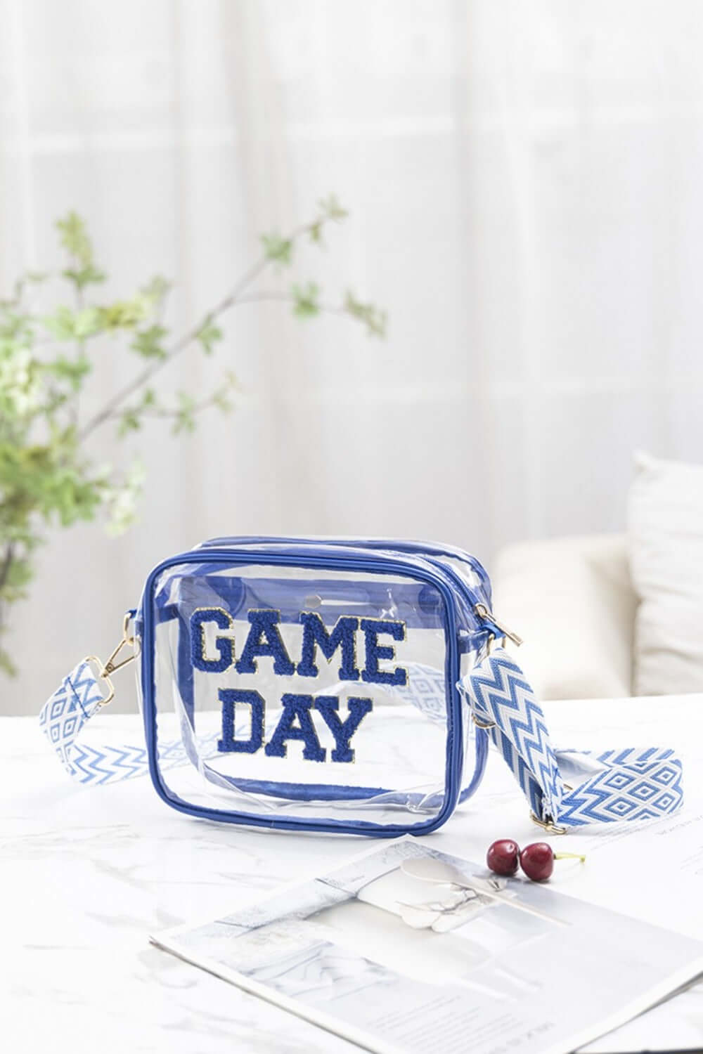 Transparent crossbody bag with "GAME DAY" letters, blue glitter trim, and zigzag strap on a white chair. Perfect for stadium events.