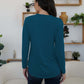 Woman wearing FAM-FAM Round Neck Long Sleeve T-Shirt in teal, seen from behind, standing in a stylish room with plants and decor.