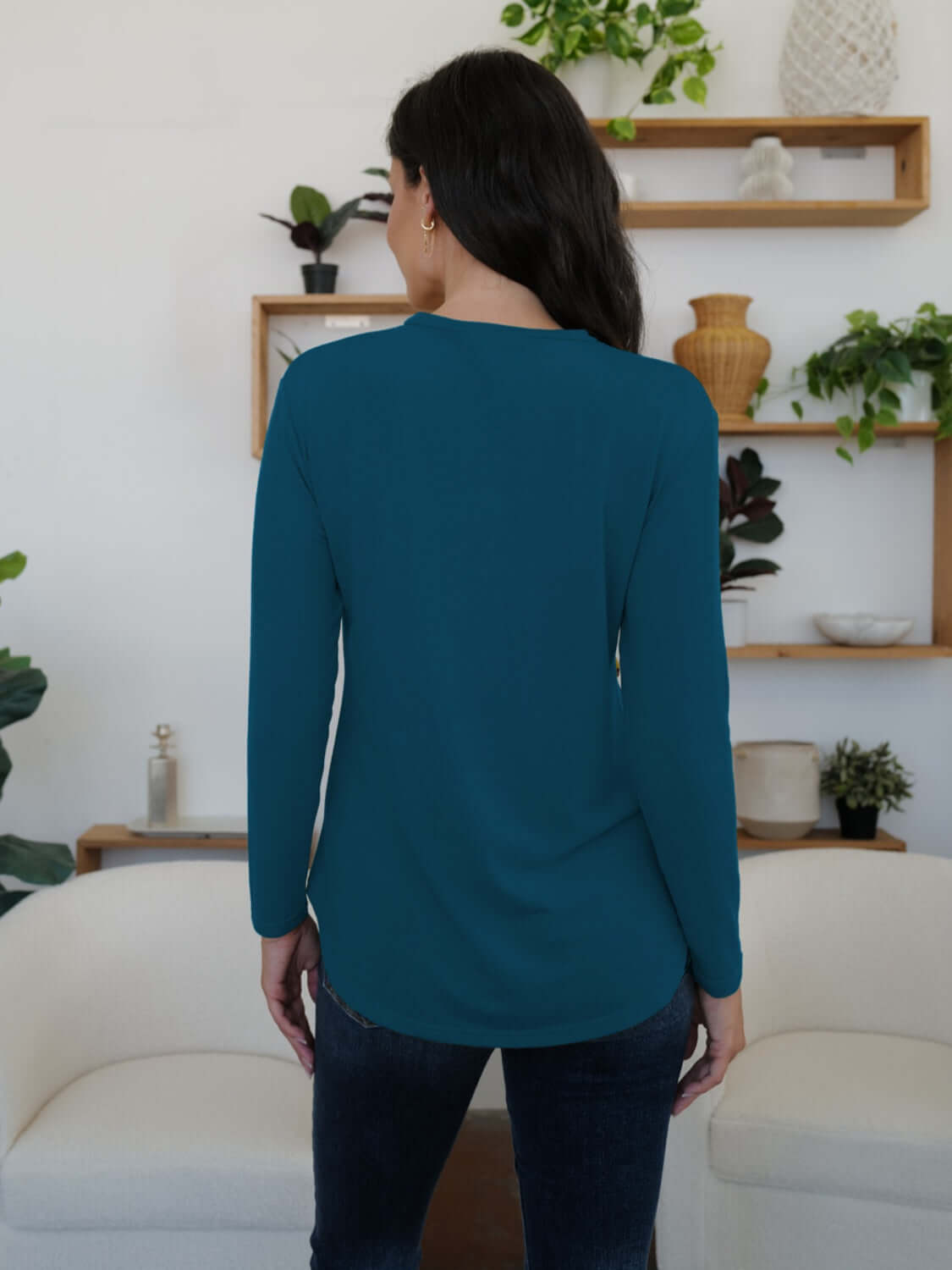 Woman wearing FAM-FAM Round Neck Long Sleeve T-Shirt in teal, seen from behind, standing in a stylish room with plants and decor.