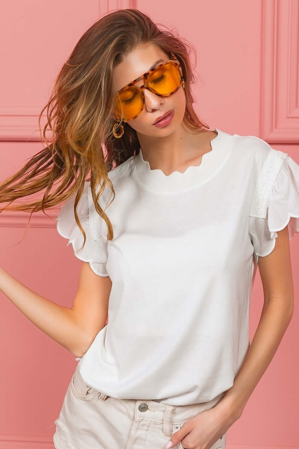 Woman wearing scallop edged lace trim top in white with intricate lace detailing, posing against a pink background