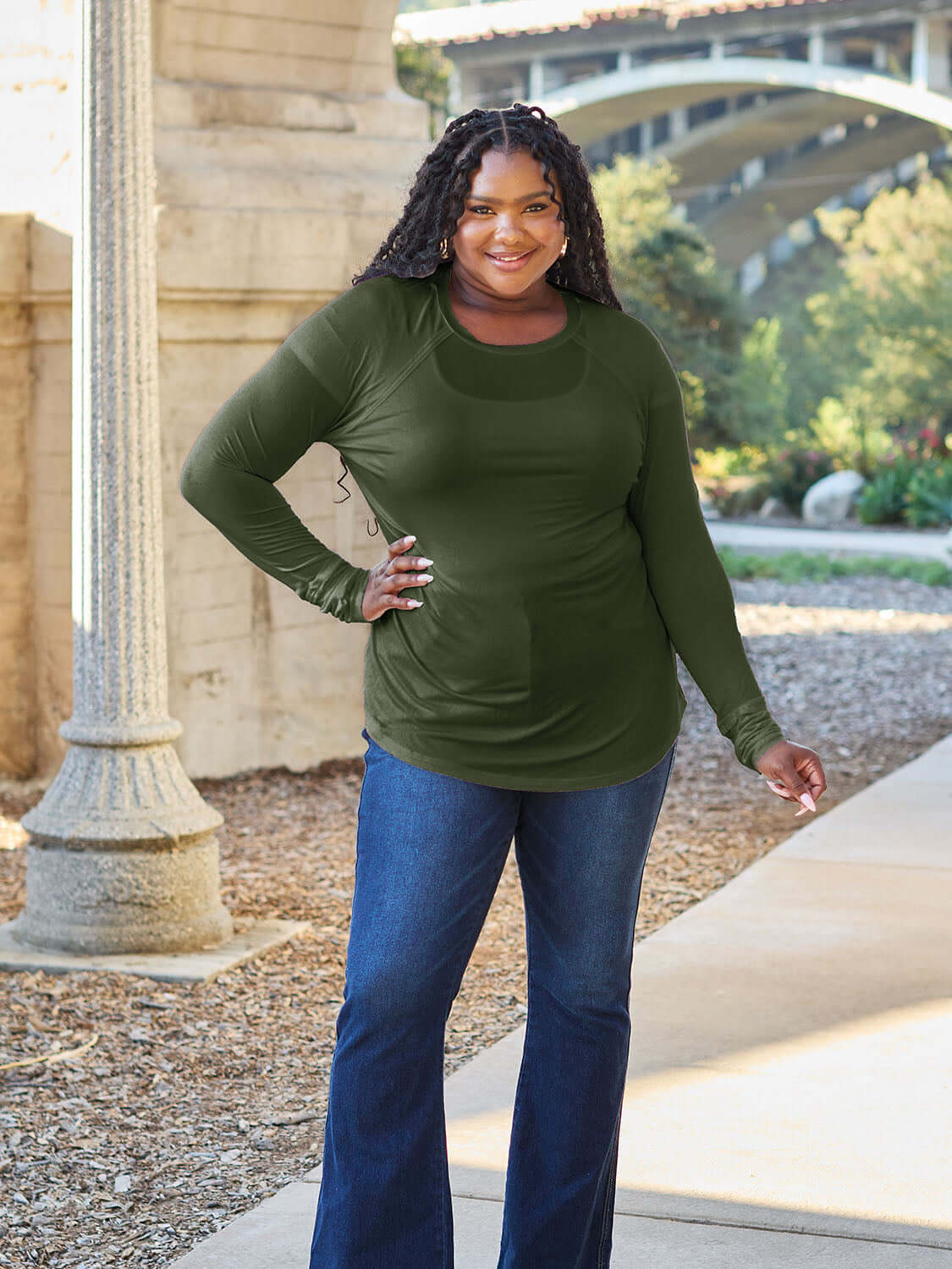 Plus-size model wearing green round neck long sleeve t-shirt, standing outdoors in a casual setting.