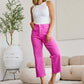 Woman wearing Crop Chloe full size tummy control high waist raw hem jeans in vibrant pink, paired with a white tank top and heels in a modern living room.