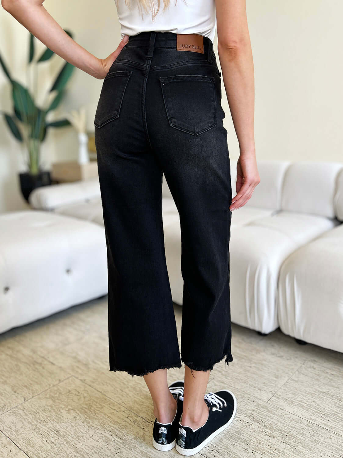 Woman wearing high waist black cropped jeans with raw hem, showcasing Judy Blue Jeans label, standing indoors near white couch.