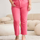 Woman wearing pink RFM Jeans Crop Dylan Full Size Tummy Control High Waist Raw Hem Jeans, standing in a stylish living room.