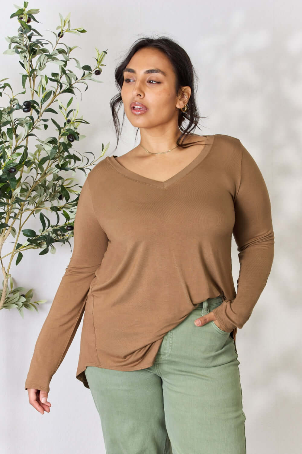 Woman wearing a long sleeve V-neck top in brown with green pants, standing near a plant.