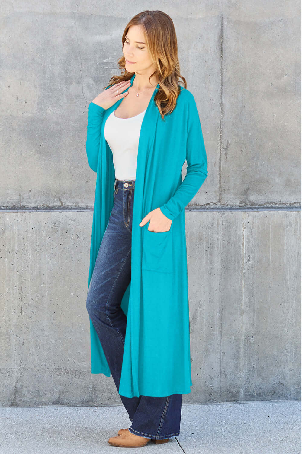 Woman wearing open front long sleeve teal cover-up with pockets, paired with dark jeans and a white top, standing against a concrete wall.