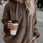 Stylish woman in a cozy brown quarter-zip drawstring teddy hoodie, holding a coffee cup on a city street.