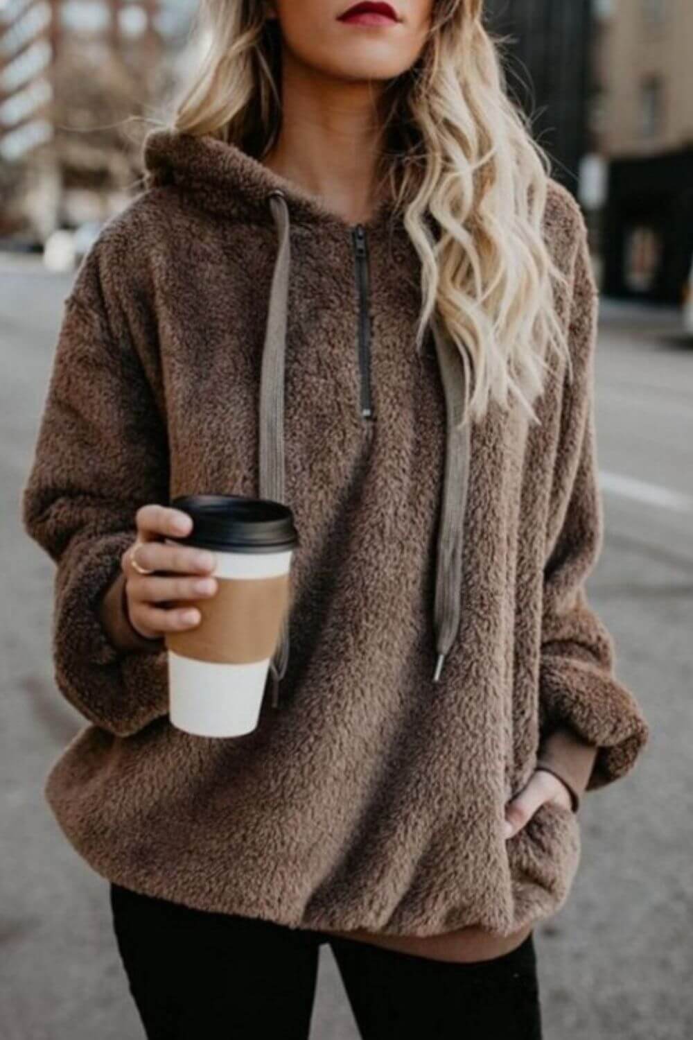 Stylish woman in a cozy brown quarter-zip drawstring teddy hoodie, holding a coffee cup on a city street.