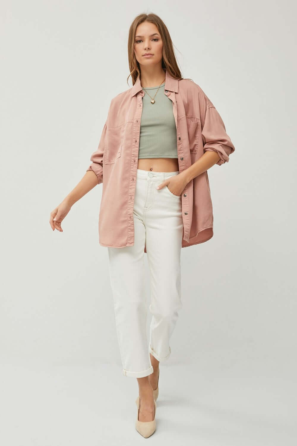 Model wearing a button-up long-sleeve pink shirt over a green crop top paired with white pants and heels.