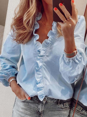 Woman wearing a light blue ruffled V-neck long sleeve blouse made of 100% polyester, styled with jeans.