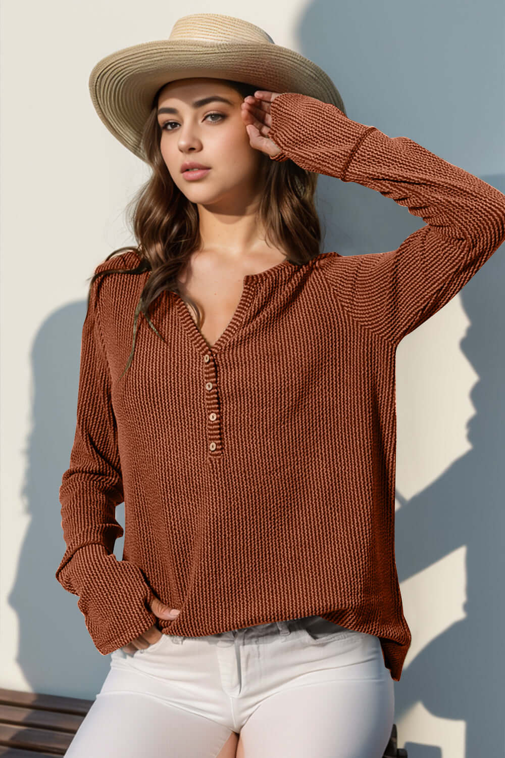 Woman in a brown notched thumbhole long sleeve t-shirt and white pants, wearing a straw hat, posing outdoors in sunlight.