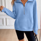 Woman wearing Ivy Lane Half Zip Raglan Sleeve Sweatshirt in light blue with pocket detail, styled with black shorts and handbag indoors.