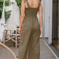 Woman wearing bodice smocked sleeveless jumpsuit in olive green, standing on a patio.