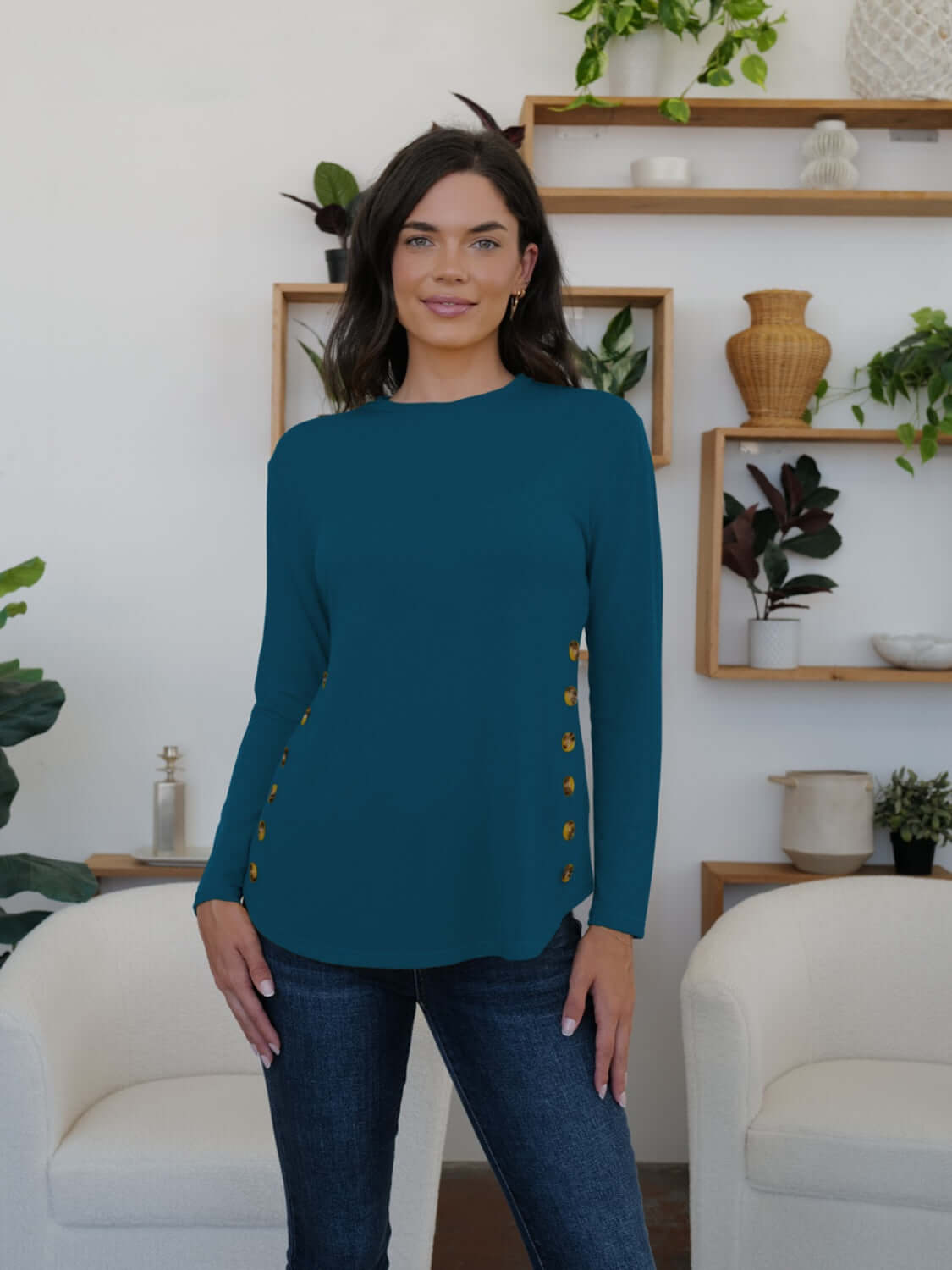 Woman in teal FAM-FAM round neck long sleeve t-shirt with decorative buttons, standing in a stylish room with plants and shelves.