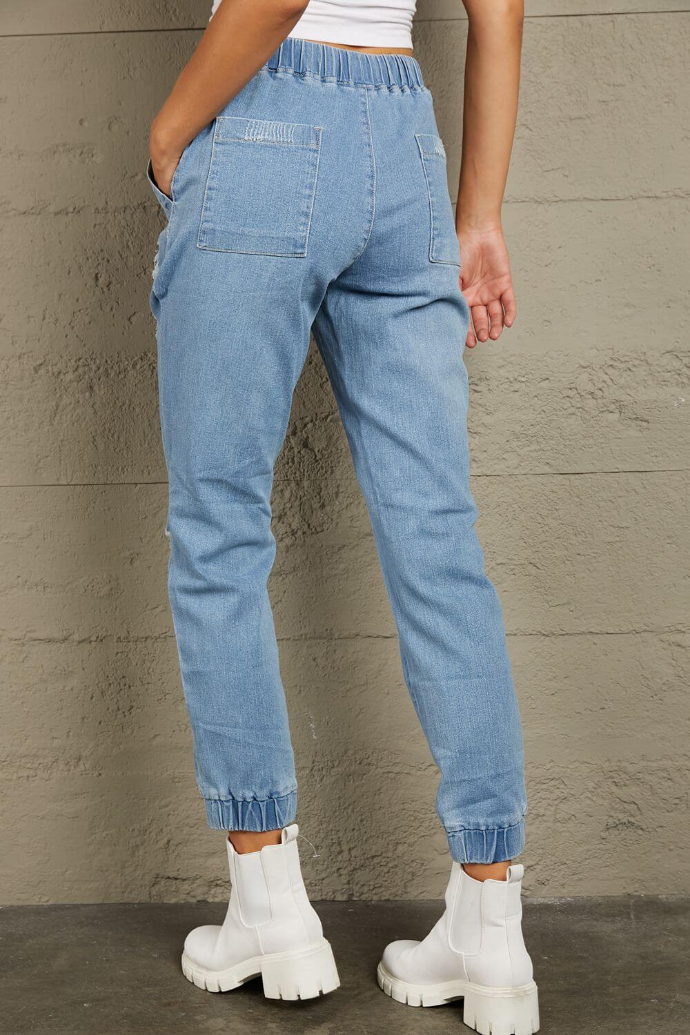 Back view of Baeful Distressed Denim Pocketed Joggers in light blue, featuring pockets and fringe, worn with white ankle boots.