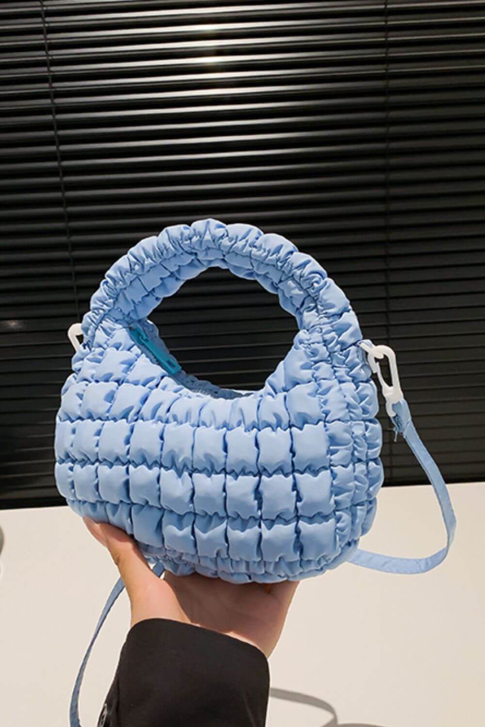 Blue quilted puffy crossbody bag with removable strap held in hand against a dark background.