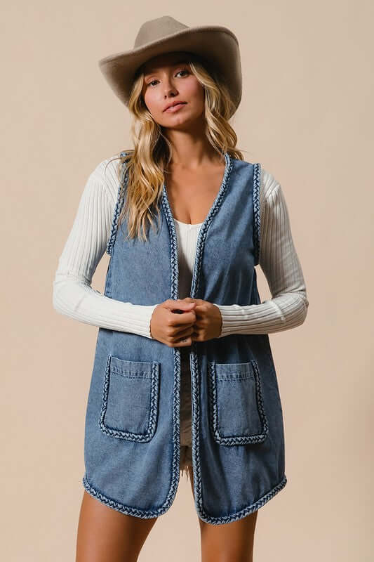 Woman wearing a stylish BiBi braided trim open front denim vest with pockets, complemented by a hat against a neutral background.