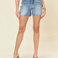 Model wearing High Waist Raw Hem Denim Shorts by Judy Blue Jeans, styled with a casual top and heels.