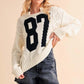 Cozy Aemi+Co cable knit sweater with contrasting number 87, styled with a casual skirt and baseball cap.