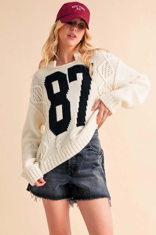 Cozy Aemi+Co cable knit sweater with contrasting number 87, styled with a casual skirt and baseball cap.