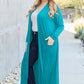 Plus-size model in teal open front long sleeve cover up with pockets, wearing sunglasses and jeans, walking on outdoor path