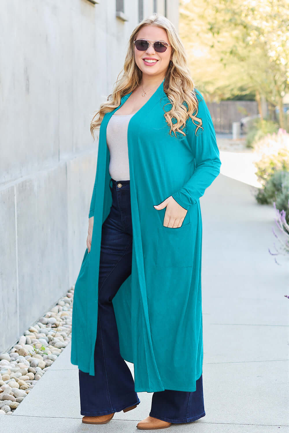 Plus-size model in teal open front long sleeve cover up with pockets, wearing sunglasses and jeans, walking on outdoor path