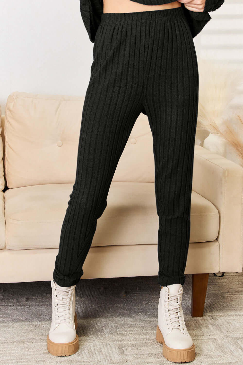 Woman wearing black ribbed pants from the Notched Long Sleeve Top and Pants Set, standing in front of beige sofa.