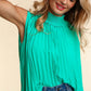 Woman wearing a green smocked mock neck pleated sleeveless top paired with blue jeans, smiling and posing with one hand in her hair.